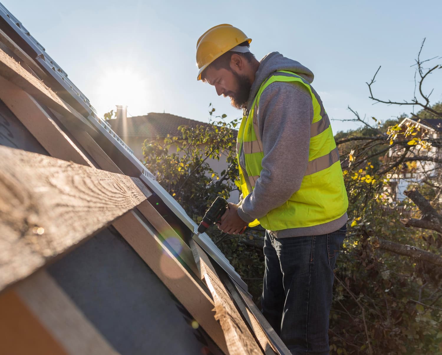 Construction worker in a yellow safety vest and helmet using a power drill to install metal roofing sheets at sunset.