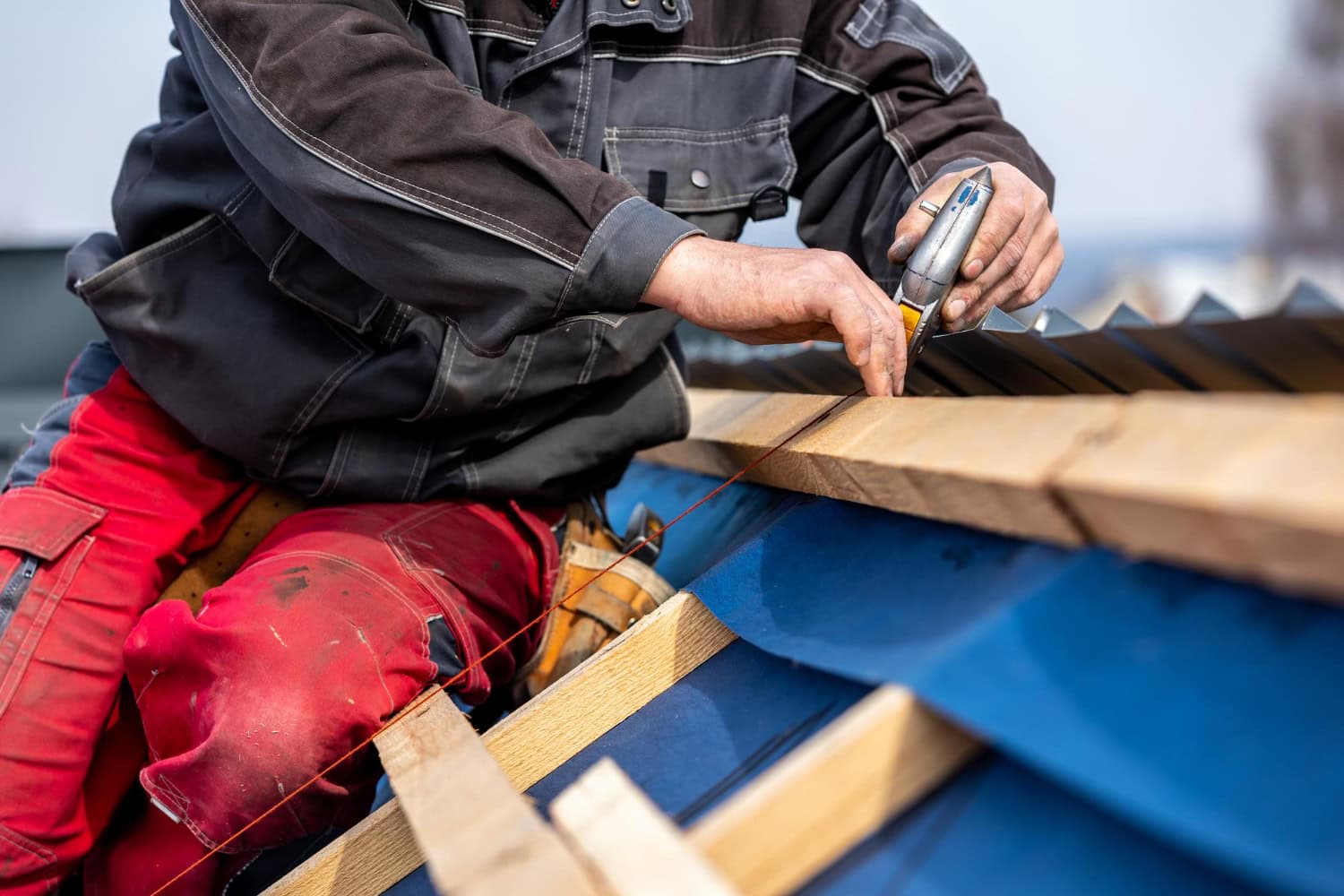 Close-up of a roofer using a utility knife and chalk line to mark and cut materials on a blue metal roof frame, wearing red work pants and a black jacket.